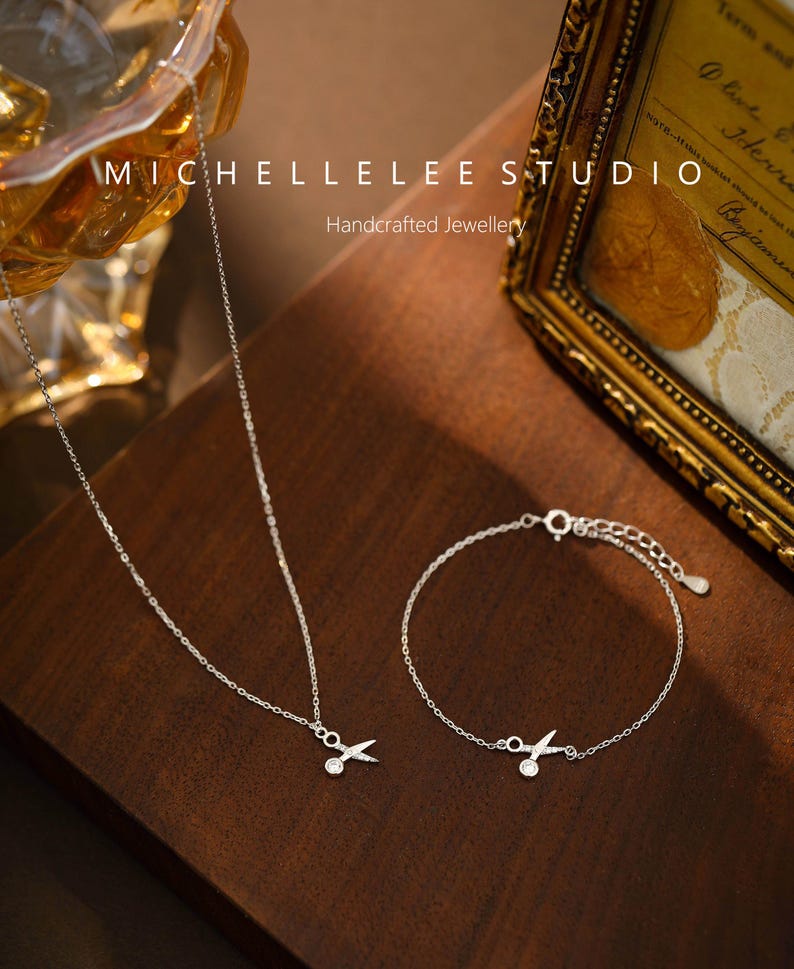 May include: A silver necklace and bracelet with a small scissor charm. The jewelry is on a wooden surface. The text "MICHELLELEE STUDIO Handcrafted Jewellery" is visible in the background.