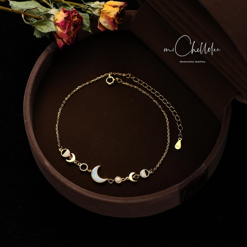 Peut inclure: Bracelet en or avec une cha&icirc;ne d&eacute;licate et cinq pendentifs en forme de croissant de lune. Chaque pendentif est orn&eacute; d'une opale blanche et d'un d&eacute;tail en &eacute;mail noir. Le bracelet est pr&eacute;sent&eacute; dans un &eacute;crin en velours marron avec l'inscription "mi Chellelee Handmade Jewelry".