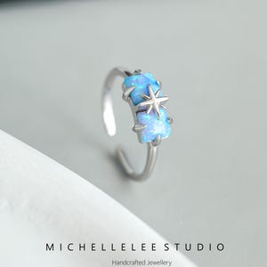 Può includere: Un anello d'argento con una pietra di opale blu e un design a stella. L'anello è su una superficie bianca. Michelle Lee Studio Gioielli artigianali.
