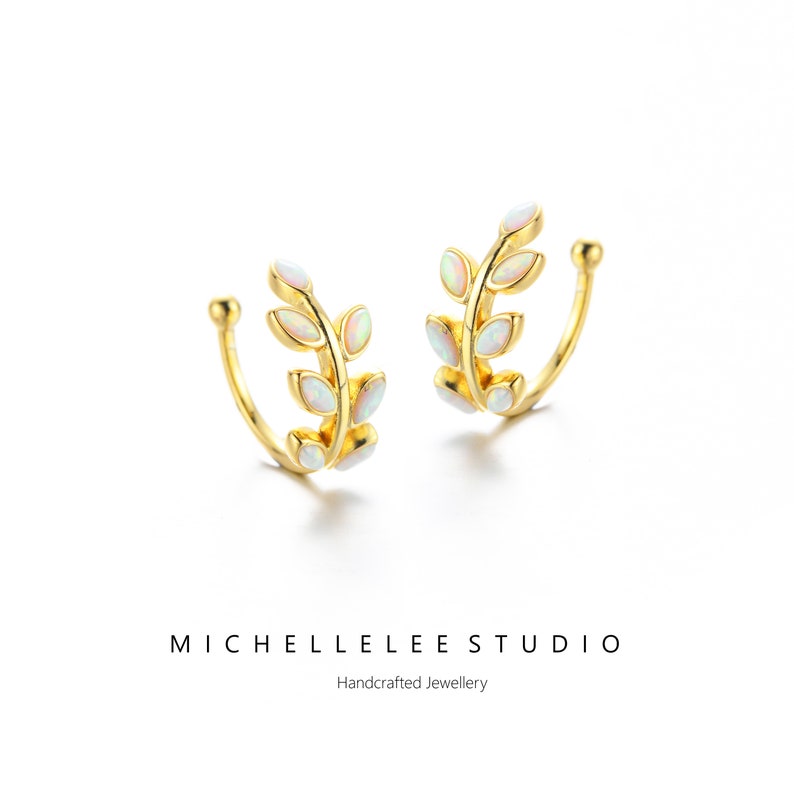 Puede incluir: Un par de pendientes tipo ear cuff dorados con un dise&ntilde;o de hojas adornado con peque&ntilde;as &oacute;palos ovalados iridiscentes. La joyer&iacute;a se presenta con el texto MICHELLELEE STUDIO Handcrafted Jewellery debajo de los objetos.