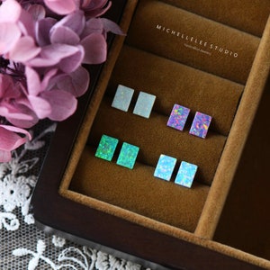 Peut inclure: Une boîte en bois avec un revêtement en velours présente six paires de boucles d'oreilles carrées en opale de différentes couleurs, notamment blanc, bleu, vert et violet. La boîte porte l'inscription "MICHELLELEE STUDIO Bijoux artisanaux" sur le dessus.