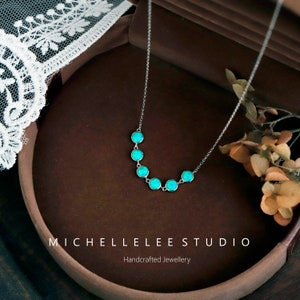 May include: A delicate silver necklace featuring seven round, turquoise-colored gemstones. The necklace is displayed on a brown surface, with the text "MICHELLELEE STUDIO Handcrafted Jewellery" visible at the bottom.