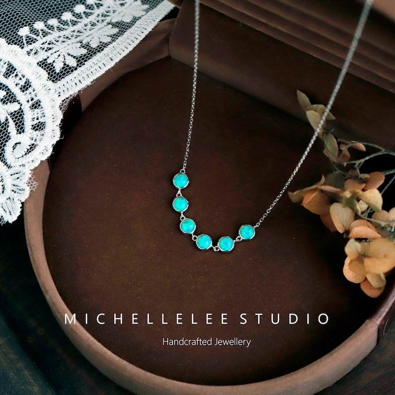 May include: A delicate silver necklace featuring seven round, turquoise-colored gemstones. The necklace is displayed on a brown surface, with the text "MICHELLELEE STUDIO Handcrafted Jewellery" visible at the bottom.