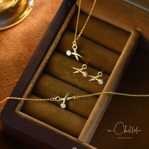 May include: A set of gold-toned jewelry featuring a necklace, earrings, and a bracelet. Each piece has a delicate design of scissors with a small clear stone in the center. The jewelry is displayed in a wooden box with the text "mi Chellee handcrafted jewelry".