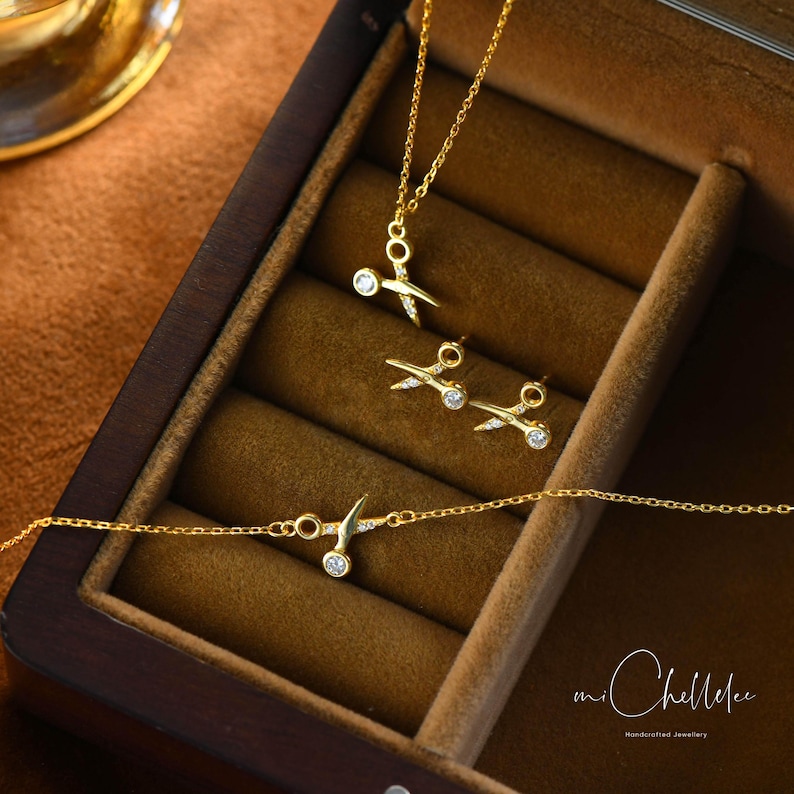 May include: A set of gold-toned jewelry featuring a necklace, earrings, and a bracelet. Each piece has a delicate design of scissors with a small clear stone in the center. The jewelry is displayed in a wooden box with the text "mi Chellee handcrafted jewelry".
