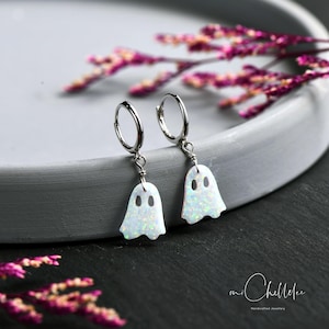 May include: A pair of silver hoop earrings featuring dangling ghost-shaped charms. The charms are made of a white, iridescent material with a shimmering, multi-colored flecked texture. Each ghost has two cut-out eye holes.