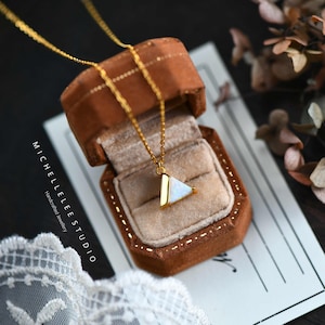 Peut inclure: Un collier en chaîne dorée avec un pendentif triangulaire en opale blanche. Le pendentif est serti dans un cadre doré et présente un design délicat. Le collier est dans une petite boîte brune avec une étiquette blanche qui indique "MICHELLELEE STUDIO Bijoux artisanaux".