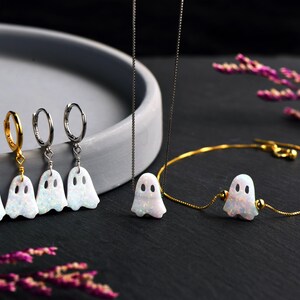 May include: A set of three gold hoop earrings and a gold necklace with a white opal ghost charm. The necklace is on a silver chain. The earrings and necklace are on a gray surface.