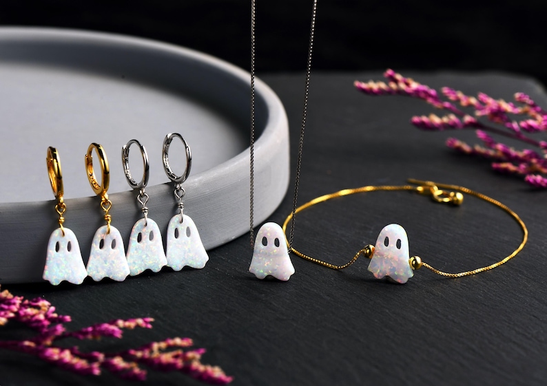 May include: A set of three gold hoop earrings and a gold necklace with a white opal ghost charm. The necklace is on a silver chain. The earrings and necklace are on a gray surface.