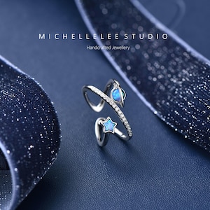 May include: A silver-toned open ring featuring a band encrusted with small clear stones, accented by a blue opal-style planet and a matching blue opal-style star. The text MICHELLELEE STUDIO Handcrafted Jewellery is visible above the ring.