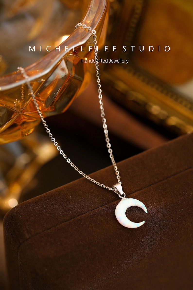 K&ouml;nnte beinhalten: Eine silberne Kettenkette mit einem Halbmond-Anh&auml;nger mit einem wei&szlig;en Opal-Stein. Die Kette wird auf einem braunen Samt-Untergrund pr&auml;sentiert. Der Text "MICHELLE LEE STUDIO" und "Handcrafted Jewellery" ist im Hintergrund sichtbar.