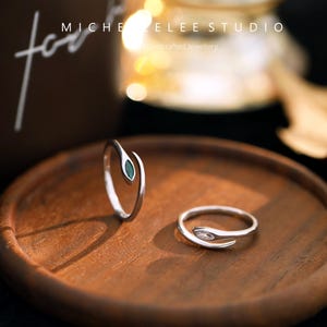 May include: Two silver rings in a snake design, one with a green stone and the other with a clear stone. The rings are presented on a wooden tray. The text "MICHELEE STUDIO" and "Handcrafted Jewellery" are visible in the background.