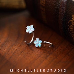 May include: Two silver flower earrings with iridescent opal stones. The earrings are in the shape of a flower with five petals. The earrings are on a wooden surface.