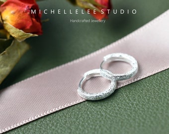 Puede incluir: Un par de pequeños pendientes de aro de plata con un acabado texturizado y brillante. Los pendientes están colocados sobre una cinta de color rosa claro. El texto MICHELLELEE STUDIO Handcrafted Jewellery es visible en la parte superior.