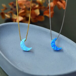 May include: Two necklaces with crescent moon pendants. One has a gold chain, the other a silver chain. The moon pendants are a light blue color with a speckled pattern. The necklaces are displayed on a light blue surface.