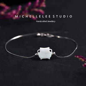 May include: A delicate silver chain bracelet with a white opal charm in the shape of a small pot. The bracelet is on a black background.  MICHELLELEE STUDIO Handcrafted Jewellery