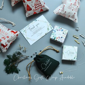 May include: A variety of handmade jewelry items are displayed on a light gray surface. The jewelry is packaged in small boxes and pouches. The boxes are decorated with colorful gemstones and the pouches are green. The jewelry includes earrings, a necklace, and a ring. The image also includes a thank you card and several small gift bags with a Christmas theme.