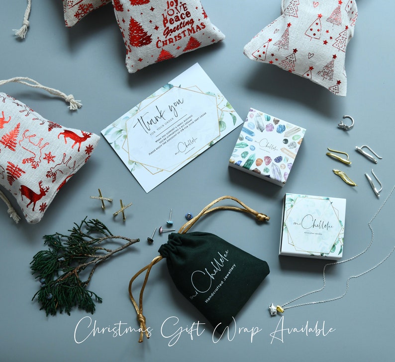May include: A variety of handmade jewelry items are displayed on a light gray surface. The jewelry is packaged in small boxes and pouches. The boxes are decorated with colorful gemstones and the pouches are green. The jewelry includes earrings, a necklace, and a ring. The image also includes a thank you card and several small gift bags with a Christmas theme.
