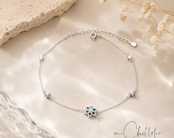 May include: A silver anklet with a floral pendant and small, sparkling details. The central flower has a light blue gemstone. The anklet is displayed on a neutral surface. The brand name "mi Chellelee" and the words "Handcrafted Jewellery" are visible.