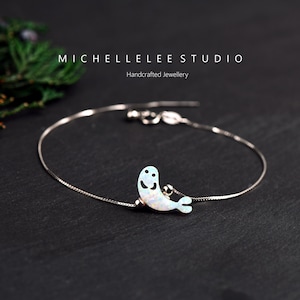 Könnte beinhalten: Ein silbernes Kettenarmband mit einem weißen Opal-Robben-Anhänger. Der Anhänger hat ein lächelndes Gesicht und ist an einer silbernen Kette befestigt. Das Armband liegt auf einem schwarzen Hintergrund. MICHELLELEE STUDIO Handgefertigte Schmuckstücke.
