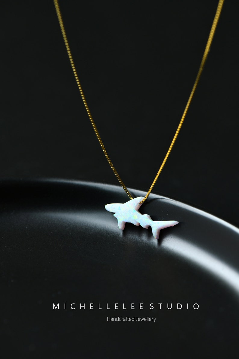 May include: A delicate gold chain necklace with a white opal shark pendant. The pendant is a simple silhouette of a shark with a shimmering opal finish. The necklace is on a black background with the text "MICHELLELEE STUDIO Handcrafted Jewellery".