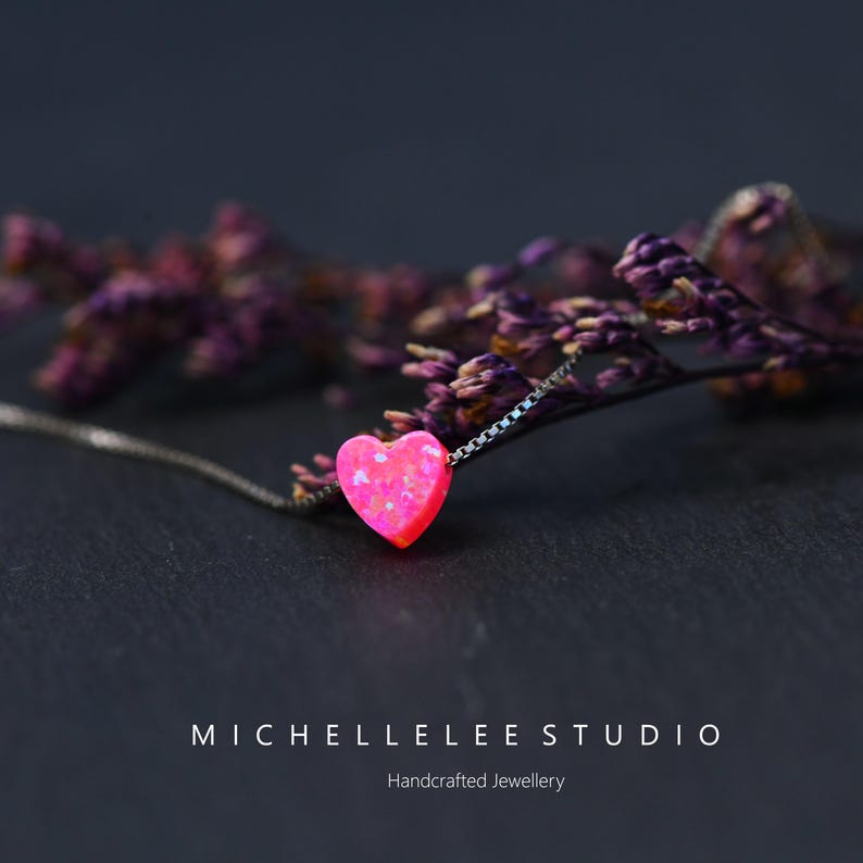 May include: A silver necklace with a bright pink heart-shaped pendant. The pendant has a textured, opal-like finish. The necklace is set against a dark backdrop with purple floral elements. The text "MICHELLELEE STUDIO Handcrafted Jewellery" is visible.