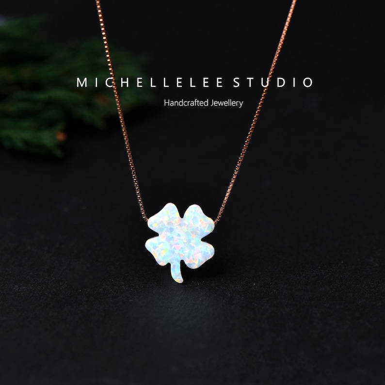 May include: A necklace with a four-leaf clover pendant. The pendant is white with iridescent flecks, hanging from a fine, rose-gold chain. The text "MICHELLELEE STUDIO Handcrafted Jewellery" is at the top.