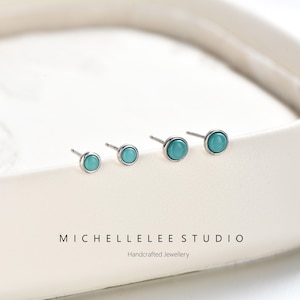 Könnte beinhalten: Vier Ohrstecker mit runden, glatten türkisfarbenen Cabochons in silberfarbenen Metallfassungen. Der Text MICHELLELEE STUDIO Handcrafted Jewellery ist unter den Ohrringen auf der cremeweißen Oberfläche aufgedruckt.