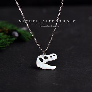 May include: A silver necklace featuring a dinosaur skull pendant. The pendant is white with a speckled design and three holes. The necklace is displayed on a black surface. The text "MICHELLE LEE STUDIO Handcrafted Jewellery" is visible.