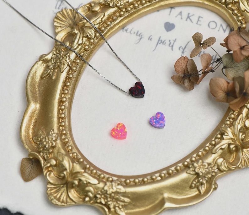 K&ouml;nnte beinhalten: Eine silberne Halskette mit herzf&ouml;rmigem Anh&auml;nger. Zwei kleine herzf&ouml;rmige Edelsteine in Rosa und Lila sind ebenfalls zu sehen. Die Artikel werden auf einer wei&szlig;en Oberfl&auml;che in einem goldfarbenen, verzierten Rahmen pr&auml;sentiert.