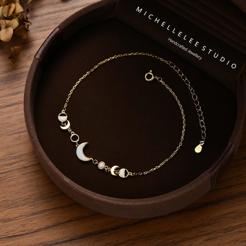 May include: A delicate gold-toned bracelet featuring a crescent moon and circular charms with white accents. The bracelet is displayed in a brown jewelry box. The text "MICHELLELEE STUDIO Handcrafted Jewellery" is visible.