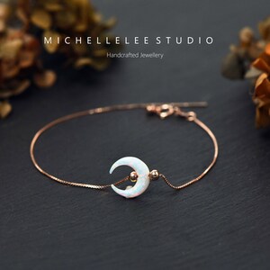 May include: A delicate rose gold chain bracelet with a crescent moon charm featuring a white opal stone. The bracelet is on a black surface with dried flowers in the background. MICHELLE LEE STUDIO Handcrafted Jewellery.