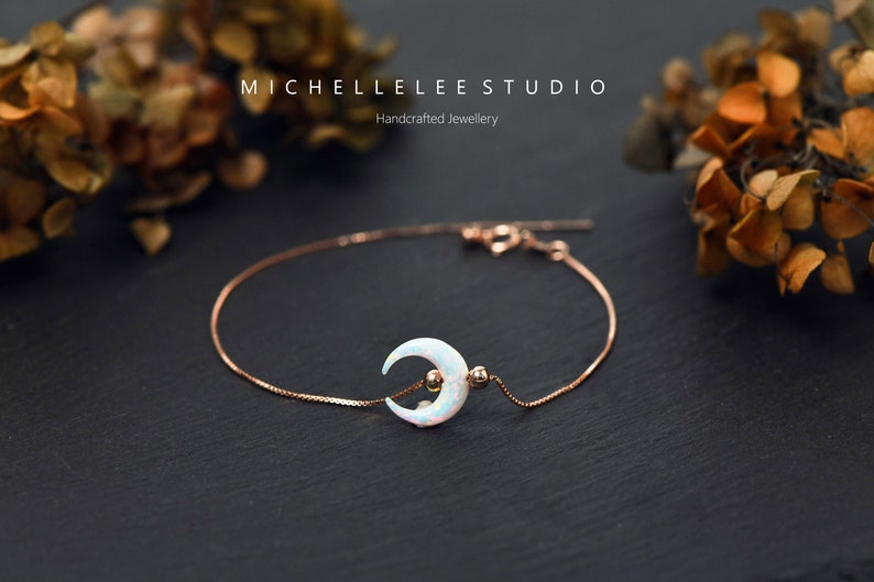 May include: A delicate rose gold chain bracelet with a crescent moon charm featuring a white opal stone. The bracelet is on a black surface with dried flowers in the background. MICHELLE LEE STUDIO Handcrafted Jewellery.