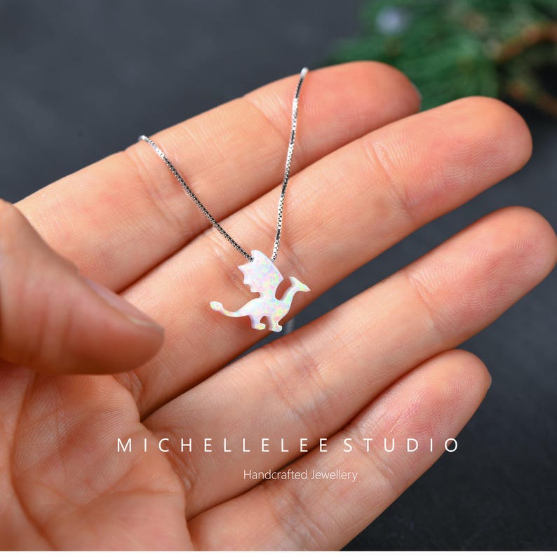 May include: A silver necklace featuring a white opal dragon pendant. The dragon has outstretched wings and a long tail. The necklace is displayed on a hand. The text "MICHELLELEE STUDIO" and "Handcrafted Jewellery" are visible.