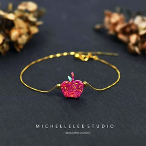 May include: A gold chain bracelet with a pink glitter apple charm. The charm has a green leaf on top. The bracelet is on a black background.