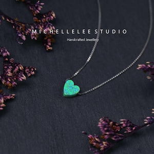 May include: A silver necklace featuring a heart-shaped pendant. The pendant is a vivid green with blue flecks. The necklace is presented against a dark backdrop, adorned with dried purple flowers. The text "MICHELLELEE STUDIO Handcrafted Jewellery" is visible.
