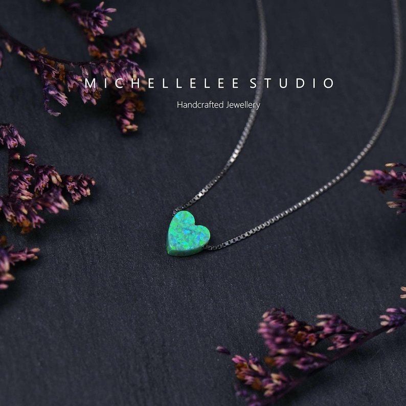 May include: A silver necklace featuring a heart-shaped pendant. The pendant is a vivid green with blue flecks. The necklace is presented against a dark backdrop, adorned with dried purple flowers. The text "MICHELLELEE STUDIO Handcrafted Jewellery" is visible.