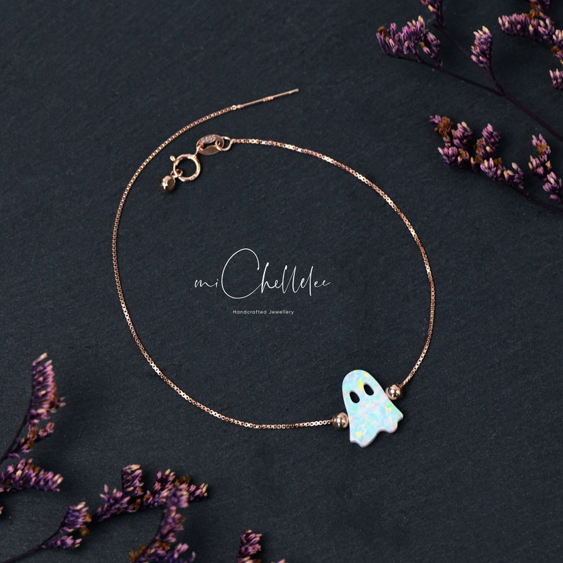 Pu&ograve; includere: Un bracciale a catena in oro rosa con un ciondolo centrale a forma di fantasma con finitura bianca iridescente. Il bracciale include una piccola chiusura a molla e due minuscole perline metalliche ai lati del fantasma. Il testo miChellelee &egrave; visibile.
