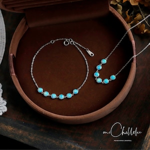 Pode incluir: Um conjunto de dois colares de prata com pedras de opala azul. O colar à esquerda é uma pulseira com seis pedras, e o colar à direita é um colar mais longo com cinco pedras. Os colares estão em uma caixa marrom com o texto "mi Chellee Handmade Jewellery".