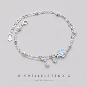 Peut inclure: Un bracelet en argent délicat avec un design à double chaîne. Orné de fleurs et de breloques en forme de goutte d'eau bleu clair, ainsi que de petites perles argentées. Le bracelet comprend le texte "MICHELLELE STUDIO" et "Handcrafted Jewellery".
