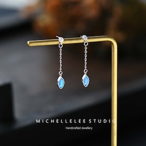 May include: A pair of silver dangle earrings featuring a blue opal stone and a small clear stone at the top. The earrings are hanging from a gold stand. The text "MICHELLELEE STUDIO Handcrafted Jewellery" is at the bottom of the image.