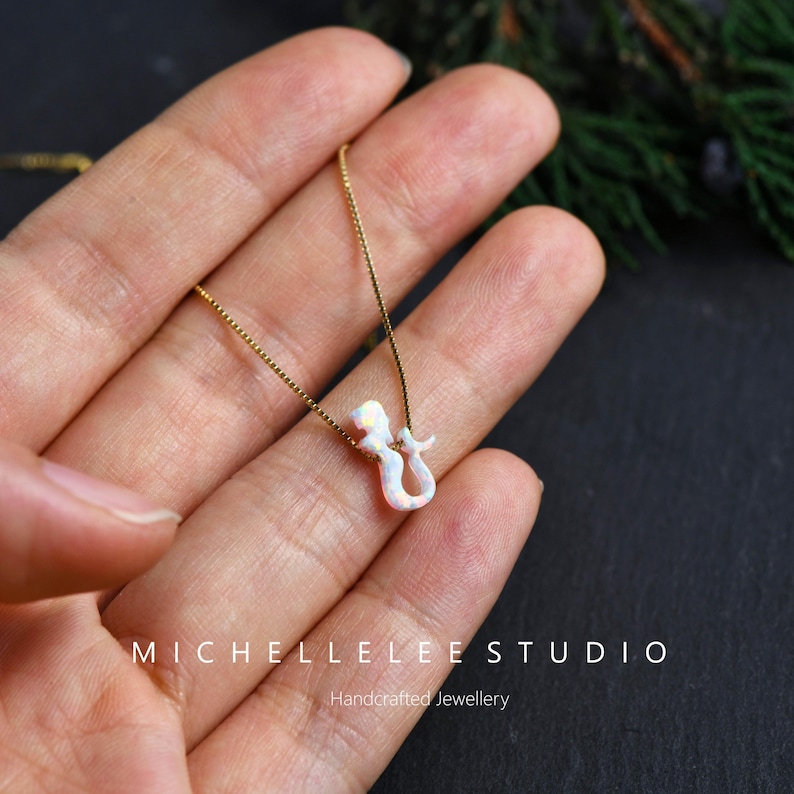 Puede incluir: Un collar de cadena dorada con un colgante de sirena de &oacute;palo blanco. El colgante tiene forma de sirena estilizada con un acabado nacarado. El collar se muestra en una mano, con el texto "MICHELLELEE STUDIO" y "Handcrafted Jewellery".