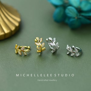 Puede incluir: Cuatro pendientes tipo ear cuff con dise&ntilde;o de hoja, dos en tono dorado y dos en tono plateado, cada uno con peque&ntilde;as piedras transparentes. El texto MICHELLELEE STUDIO Handcrafted Jewellery aparece en la parte inferior.