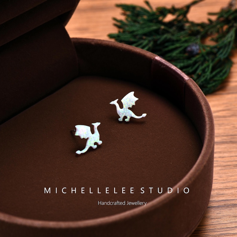 May include: A pair of opal dragon stud earrings displayed in a brown jewelry box. The earrings are white with iridescent accents, featuring a dragon design with outstretched wings. The box is open, revealing the earrings against a brown interior. The text "MICHELLELEE STUDIO" and "Handcrafted Jewellery" are visible.
