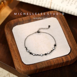 May include: A delicate black chain bracelet with white moon and star charms. The bracelet is displayed on a wooden tray with the text "MICHELLE LEE STUDIO Handcrafted Jewellery".