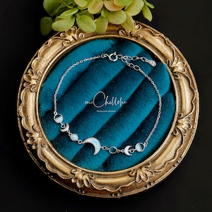 May include: A silver bracelet featuring a crescent moon and moon phase designs with opalescent accents. The bracelet is displayed on a teal velvet surface within a gold-toned ornate frame. The brand name "mi Chellelee" is visible.