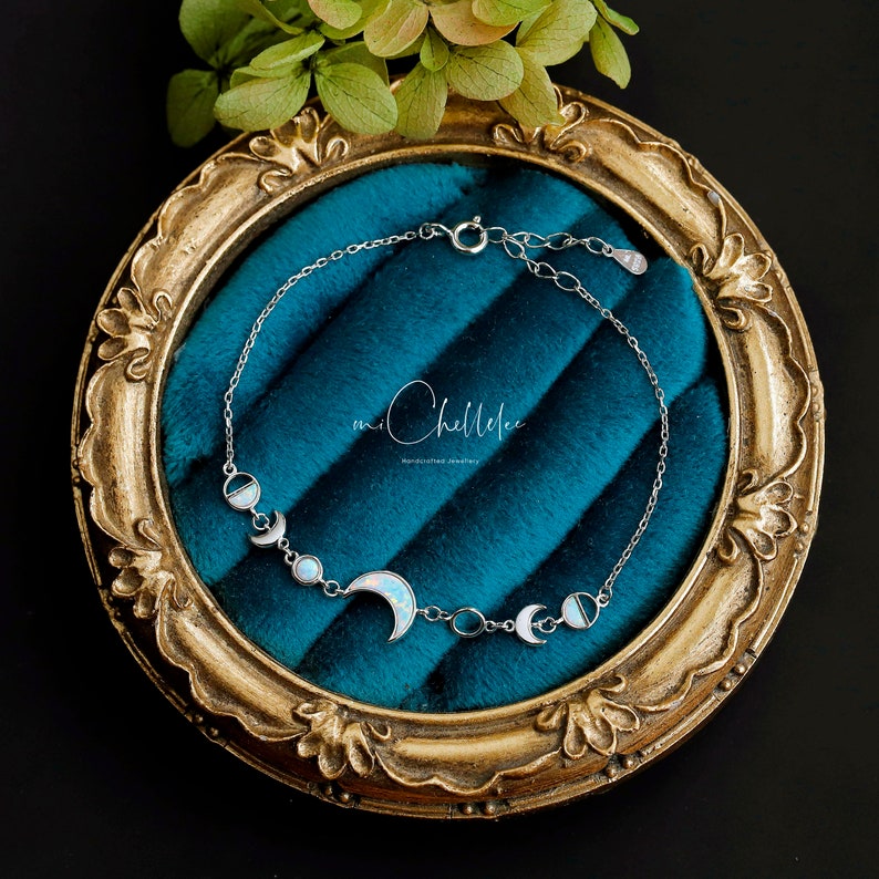 May include: A silver bracelet featuring a crescent moon and moon phase designs with opalescent accents. The bracelet is displayed on a teal velvet surface within a gold-toned ornate frame. The brand name "mi Chellelee" is visible.
