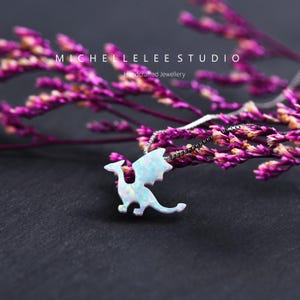 May include: A silver necklace featuring a dragon pendant with an iridescent, opal-like finish. The dragon is silhouetted against a backdrop of purple flowers and a dark surface. The text "MICHELLELEE STUDIO" and "Handcrafted Jewellery" are visible.