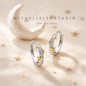 May include: A pair of silver hoop earrings featuring a row of small clear stones and gold-toned star accents. The text MICHELLELEE STUDIO Handcrafted Jewellery is visible above the earrings, along with a large crescent moon and small stars.