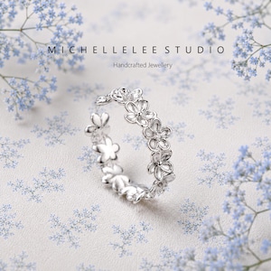 May include: A silver ring featuring a continuous band of small, five-petalled flowers. The text MICHELLELEE STUDIO Handcrafted Jewellery is visible above the ring. The ring has a polished metallic finish and a delicate, open-work floral design.